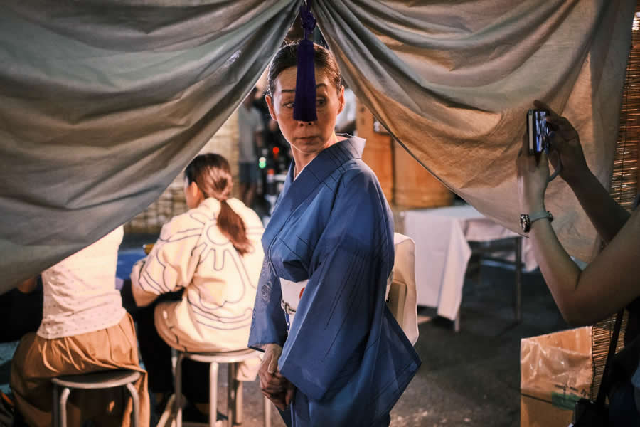 The Art of Perfect Timing: 34 Clever Street Photos by Emiliano Cuadrado That Turn Ordinary Scenes Into Visual Surprises 51 A woman wearing a blue traditional kimono glances sideways through parted curtains, framed by people, warm ambient light, and layered foreground elements in a cinematic street photography composition.
