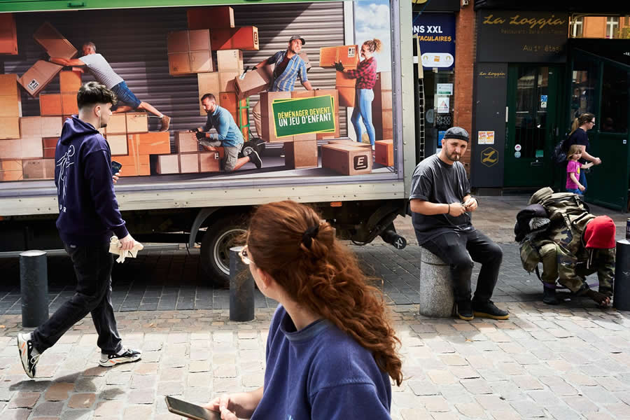 The Art of Perfect Timing: 34 Clever Street Photos by Emiliano Cuadrado That Turn Ordinary Scenes Into Visual Surprises 52 Street scene featuring pedestrians beside a delivery truck covered with an advertisement of people moving boxes, creating a playful visual connection through layered composition and perfectly timed street photography.