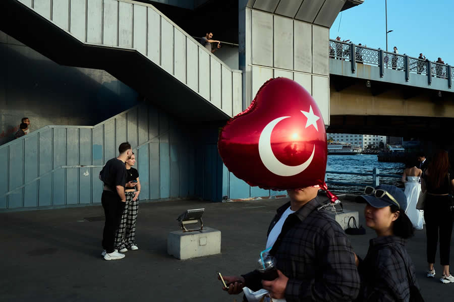 The Art of Perfect Timing: 34 Clever Street Photos by Emiliano Cuadrado That Turn Ordinary Scenes Into Visual Surprises 53 A person carrying a red heart-shaped balloon with the Turkish flag has their face completely hidden, framed against a riverside bridge and geometric architecture in a perfectly timed street photograph.