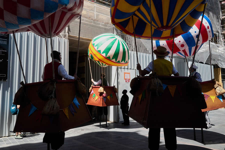 The Art of Perfect Timing: 34 Clever Street Photos by Emiliano Cuadrado That Turn Ordinary Scenes Into Visual Surprises 56 Performers wearing colorful hot-air-balloon costumes stand in a sunlit street, framed by bold patterns, silhouettes, and layered urban surroundings in a creative street photography composition.