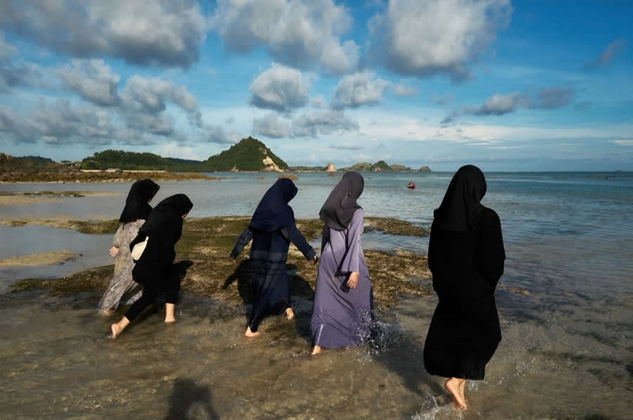 The Art of Perfect Timing: 34 Clever Street Photos by Emiliano Cuadrado That Turn Ordinary Scenes Into Visual Surprises 59 Four women in flowing dresses and headscarves walk through shallow seawater toward the horizon, framed by dramatic clouds, distant islands, and a wide coastal landscape in a serene street photography scene.