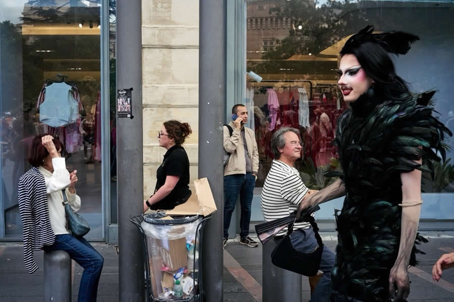 The Art of Perfect Timing: 34 Clever Street Photos by Emiliano Cuadrado That Turn Ordinary Scenes Into Visual Surprises 60 A performer wearing an elaborate black feathered costume walks past unsuspecting pedestrians outside a storefront, creating a dramatic contrast in a candid, layered street photography composition.