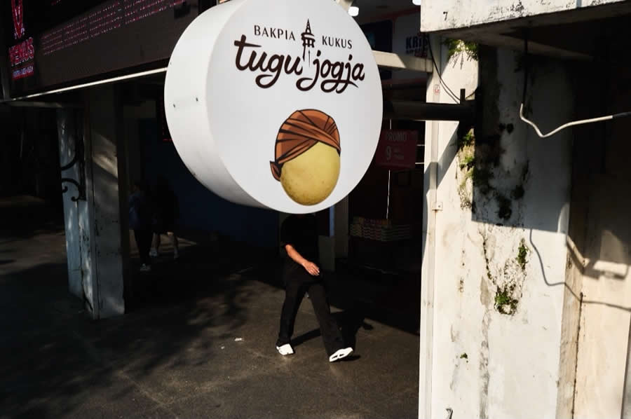 The Art of Perfect Timing: 34 Clever Street Photos by Emiliano Cuadrado That Turn Ordinary Scenes Into Visual Surprises 63 A pedestrian walking beneath a round hanging shop sign appears to have the illustrated face on the sign, creating a humorous optical illusion in a perfectly timed street photography composition.