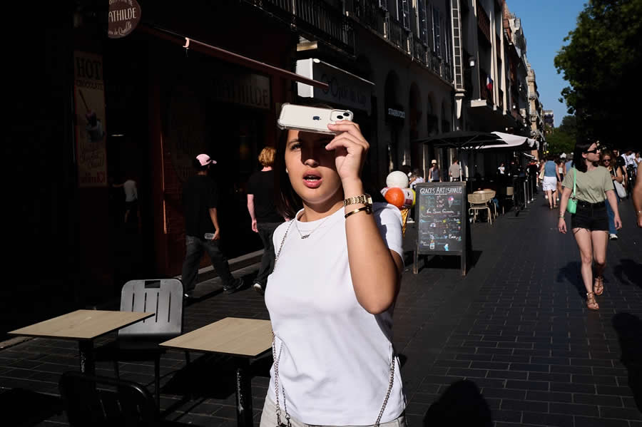 The Art of Perfect Timing: 34 Clever Street Photos by Emiliano Cuadrado That Turn Ordinary Scenes Into Visual Surprises 65 A woman shades her face with a smartphone while walking along a sunlit city street, with an orange balloon aligning behind her to create a playful, perfectly timed street photography composition.