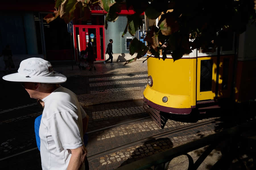 The Art of Perfect Timing: 34 Clever Street Photos by Emiliano Cuadrado That Turn Ordinary Scenes Into Visual Surprises 66 A pedestrian in a white hat walks past a bright yellow tram beneath leafy branches, framed by dramatic sunlight, shadows, and patterned cobblestone streets in a layered street photography composition.