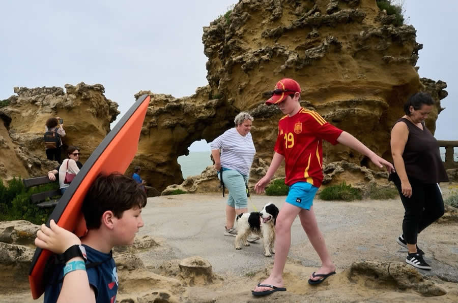 The Art of Perfect Timing: 34 Clever Street Photos by Emiliano Cuadrado That Turn Ordinary Scenes Into Visual Surprises 71 People explore rugged coastal rock formations while a boy carrying a bright orange bodyboard walks through the foreground, creating a layered street photography composition with natural arches and candid human interactions.