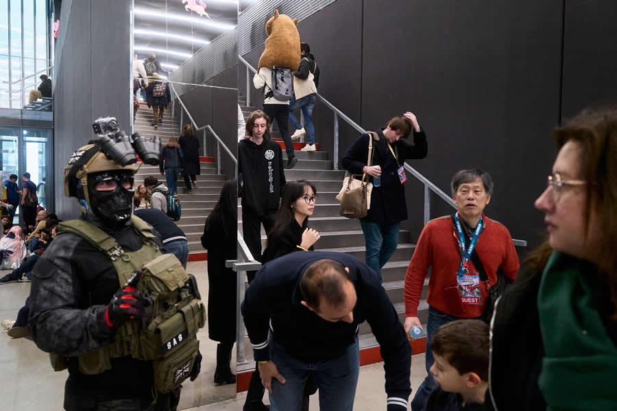 The Art of Perfect Timing: 34 Clever Street Photos by Emiliano Cuadrado That Turn Ordinary Scenes Into Visual Surprises 72 A person in tactical cosplay stands among visitors inside a convention hall, while people move across staircases and a giant bear mascot climbs above, creating a layered, dynamic street photography composition.