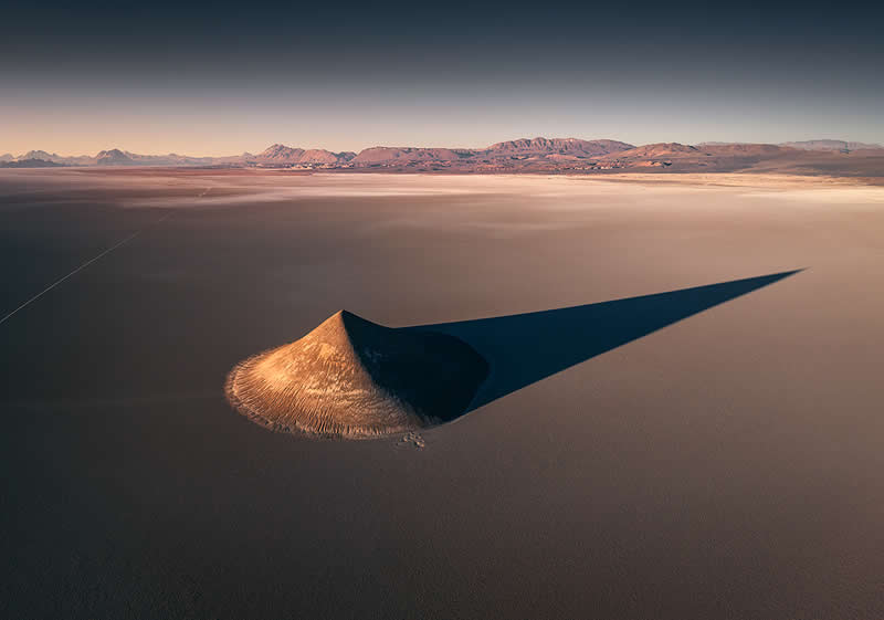 A vast Atacama Desert salt flat with a small mound in the foreground casting a long, dramatic shadow toward distant reddish mountains beneath a clear sky.