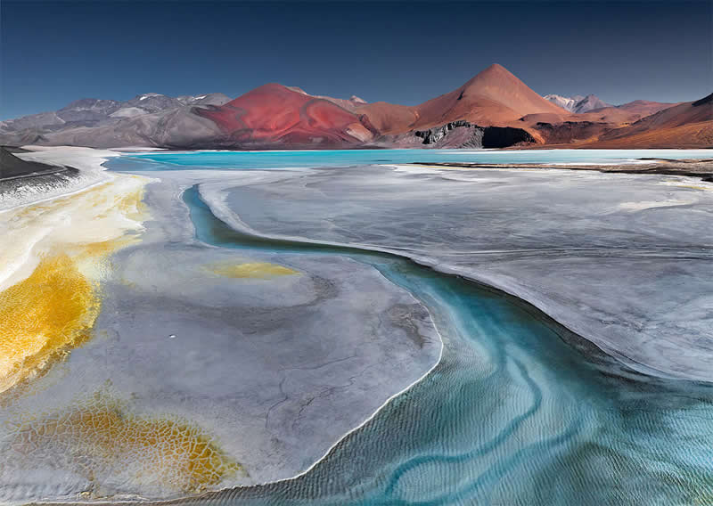 A surreal Atacama Desert landscape featuring a turquoise mineral lagoon winding through pale salt flats, golden mineral textures, and colorful red and gray mountains beneath a deep blue sky.