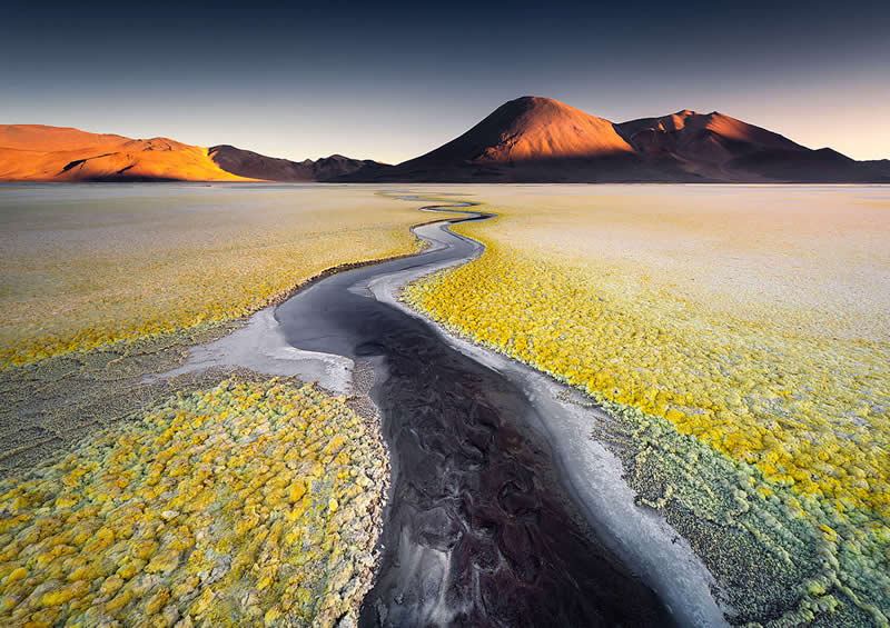 A dark winding channel cuts through golden mineral-covered ground in the Atacama Desert, leading toward dramatic volcanic mountains illuminated by warm sunlight beneath a deep blue sky.