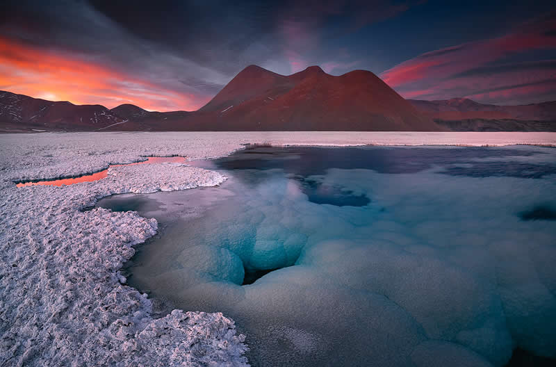 A surreal Atacama Desert landscape with a turquoise mineral lagoon surrounded by pale salt formations, dark volcanic mountains, and a vivid pink-orange sunset reflected across the water.