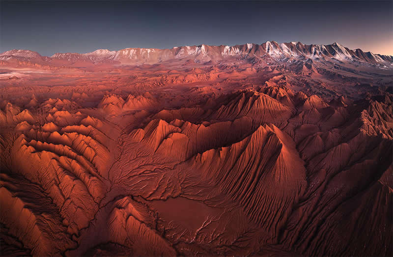 A sweeping aerial view of the Atacama Desert showing deeply eroded red-orange ridges and valleys stretching toward a rugged mountain range beneath a dark, clear sky.