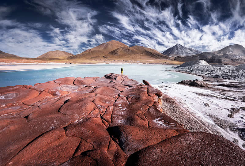 A vast Atacama Desert landscape with cracked red rock in the foreground, a turquoise lagoon, colorful volcanic mountains, dramatic clouds, and a tiny person standing near the water.