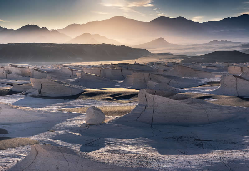 A surreal Atacama Desert landscape featuring pale, cracked rock formations in the foreground, layered mountain ranges in the distance, and soft golden sunlight illuminating the hazy terrain.