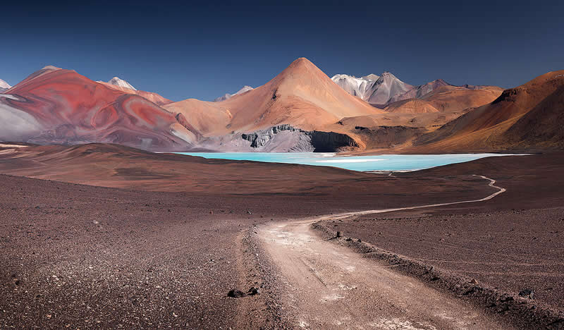 A winding dirt trail crosses the barren Atacama Desert toward a turquoise lagoon and colorful volcanic mountains beneath a deep blue sky.