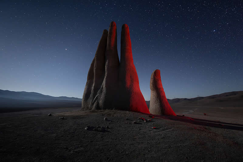 A monumental hand-shaped rock formation rises from the dark Atacama Desert beneath a star-filled night sky, dramatically illuminated with red light against distant mountains.
