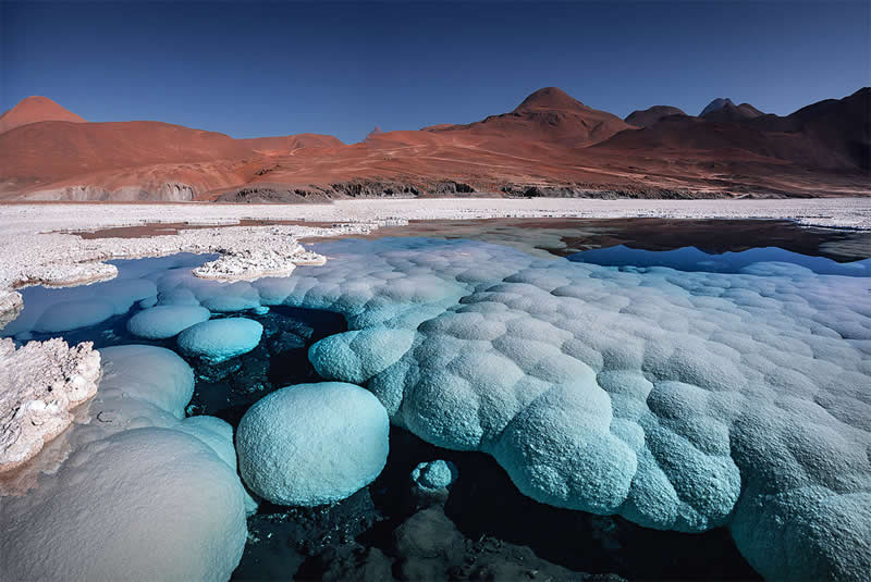 A surreal Atacama Desert landscape showing rounded pale-blue mineral formations surrounding dark turquoise water, with white salt crusts and reddish volcanic mountains beneath a deep blue sky.