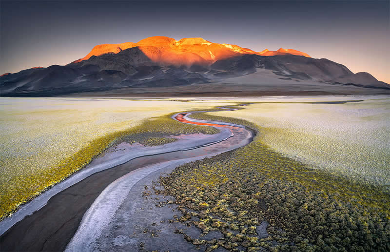 A winding dark mineral channel crosses the Atacama Desert toward a massive volcanic mountain glowing orange in sunlight, surrounded by pale mineral textures beneath a deep blue sky.