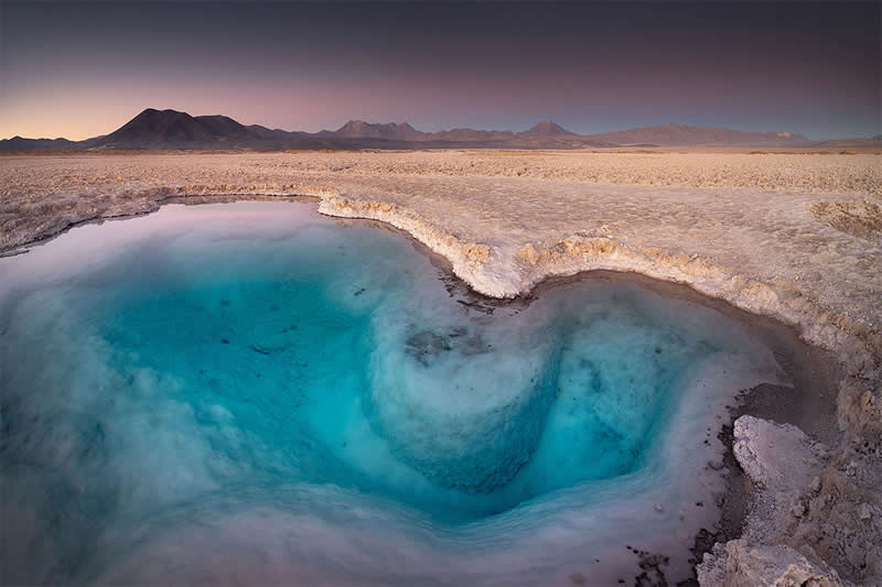 A vivid turquoise mineral lagoon curves through the pale salt flats of the Atacama Desert, with distant dark mountains beneath a softly colored evening sky.