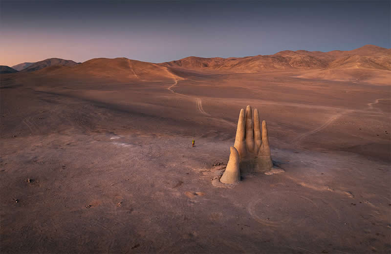A giant hand-shaped sculpture rises from the barren Atacama Desert, with a tiny person standing nearby and expansive brown hills stretching across the background beneath a muted sky.