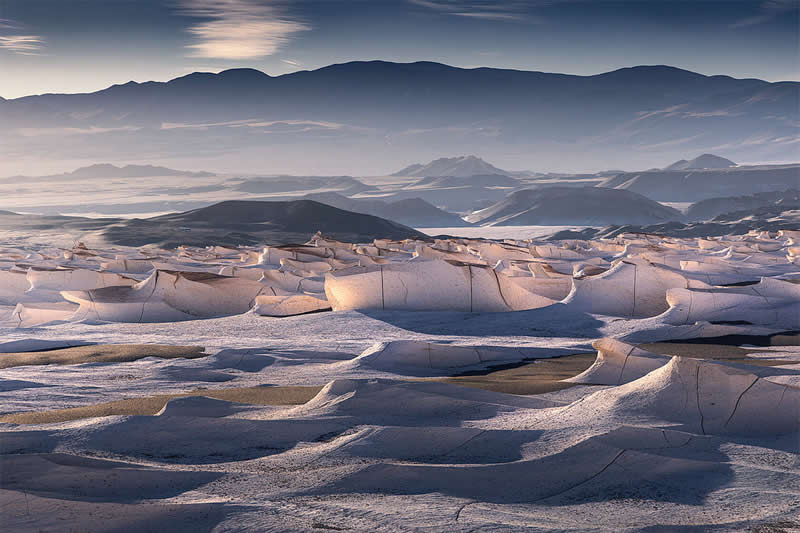 A vast Atacama Desert landscape filled with pale, wave-like mineral formations, stretching toward layered mountain ranges under a hazy blue sky and soft atmospheric light.