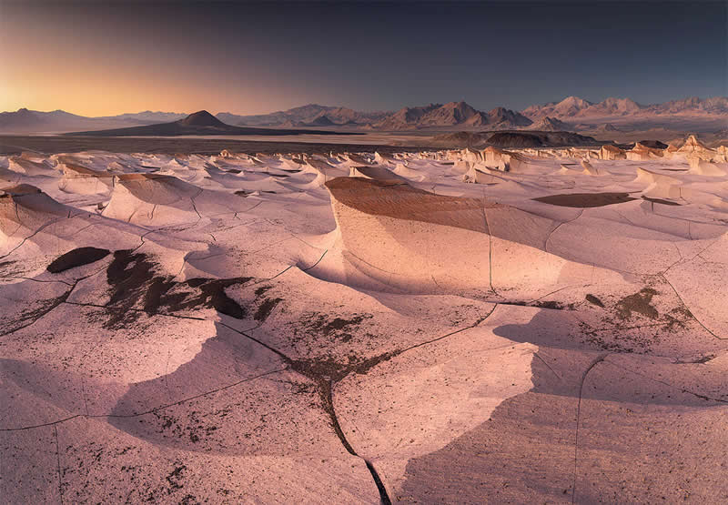 A sweeping Atacama Desert landscape with pale sculpted ridges and deep shadows stretching toward distant rugged mountains, illuminated by warm evening light beneath a darkening sky.