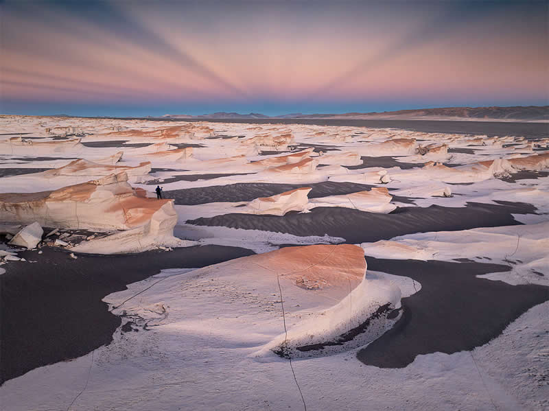 Expansive Atacama Desert landscape featuring pale sculpted rock formations surrounded by dark ground, with tiny figures in the distance beneath a soft pink and blue evening sky.