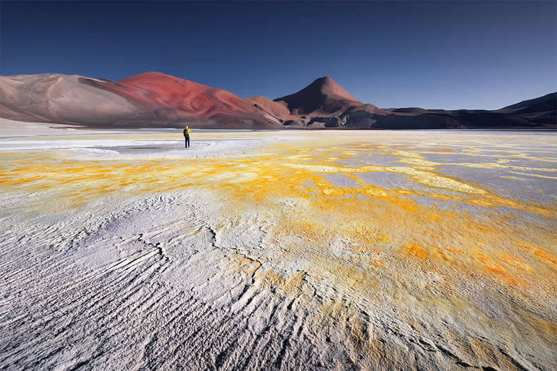 Wide Atacama Desert scene showing rippled white salt flats streaked with golden-yellow mineral textures, colorful volcanic mountains in the background, and a tiny solitary figure beneath a deep blue sky.