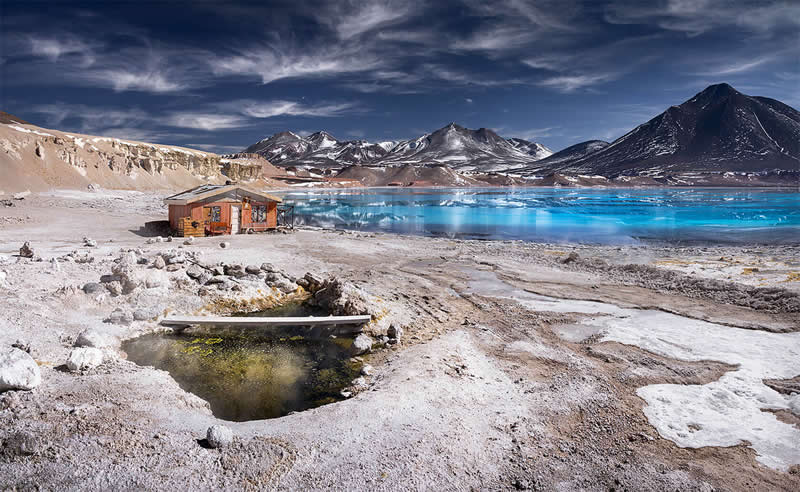 A vivid blue mineral lagoon in the Atacama Desert surrounded by snow-dusted volcanic mountains, with a small weathered wooden shelter and steaming mineral pool in the barren foreground.