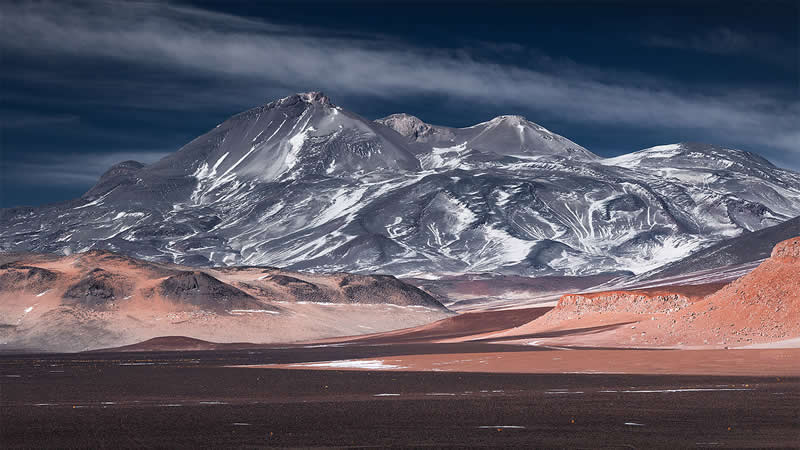 Expansive Atacama Desert landscape featuring a dark volcanic plain, reddish rocky ridges, and towering snow-streaked mountains beneath a dramatic sky filled with sweeping clouds.