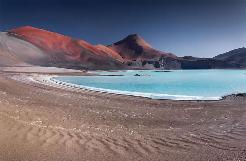 A brilliant turquoise lagoon curves through the barren Atacama Desert, surrounded by reddish volcanic mountains and textured dark sand beneath a clear deep-blue sky.