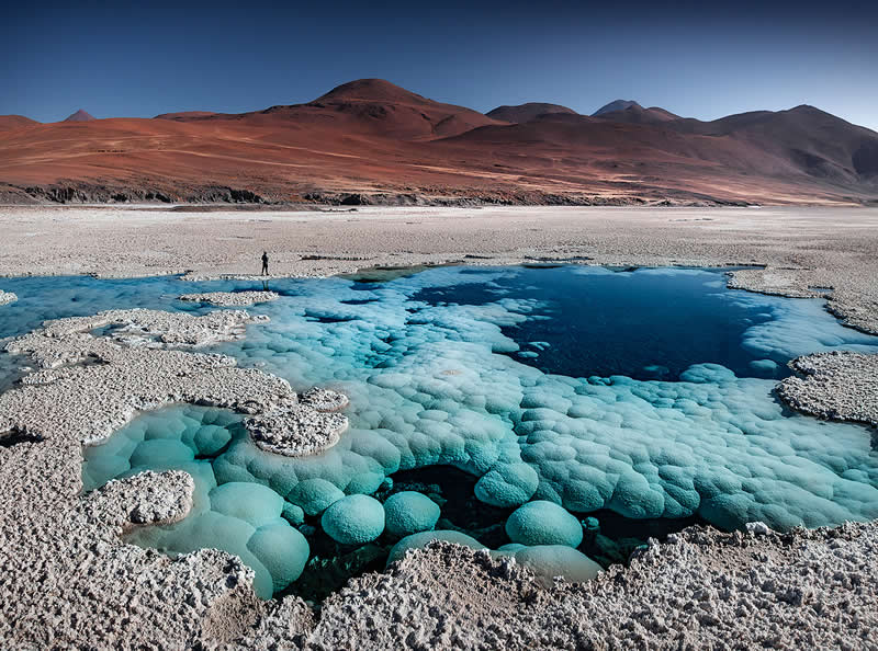 A vivid turquoise mineral pool surrounded by white salt formations in the Atacama Desert, with a tiny person standing nearby and reddish volcanic mountains stretching across the background.
