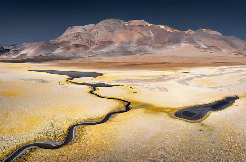 A winding dark watercourse cuts through golden and white mineral flats in the Atacama Desert, leading toward a massive multicolored mountain beneath a deep blue sky.