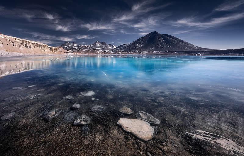 A crystal-clear turquoise lagoon reflects dark volcanic mountains and dramatic clouds in the Atacama Desert, with textured rocky shallows filling the foreground beneath a deep blue sky.