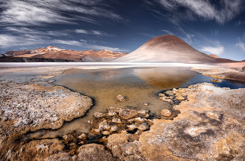 A shallow mineral lagoon reflects a smooth volcanic mountain in the Atacama Desert, surrounded by textured salt formations and rugged ground beneath a dramatic blue sky with wispy clouds.