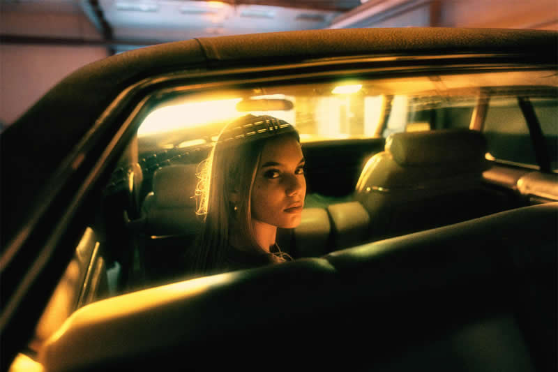 Cinematic portrait of a woman sitting inside a car, illuminated by warm golden light, with the vehicle&rsquo;s window and frame creating a dramatic, intimate composition.
