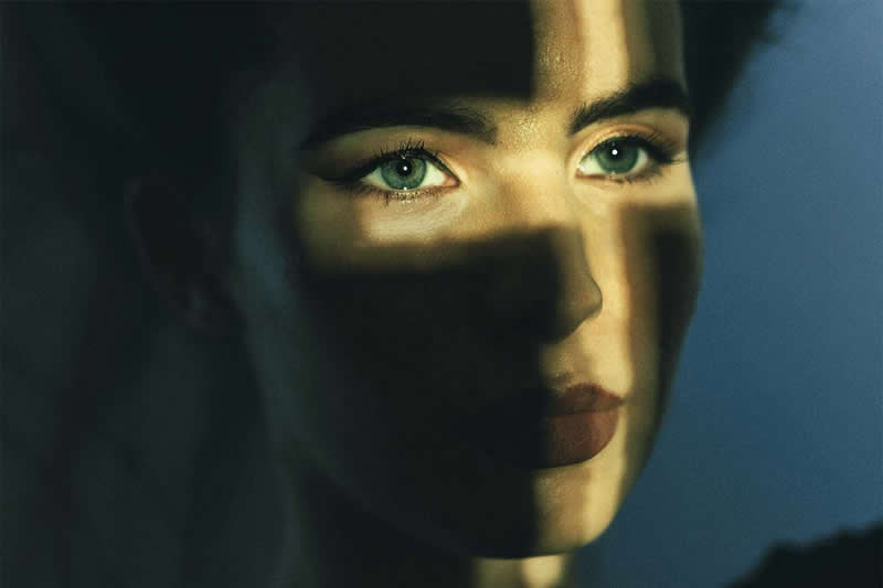 Cinematic close-up portrait of a woman&rsquo;s face partially obscured by dramatic cross-shaped shadows, with her bright eyes illuminated against a dark blue background.