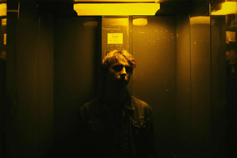 Cinematic portrait of a man standing inside a dim elevator, illuminated by dramatic golden-yellow overhead light with deep shadows surrounding his face and body.