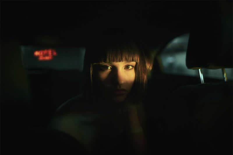 Cinematic portrait of a woman sitting inside a dark car, with warm light illuminating her eyes and part of her face while the surrounding interior fades into deep shadow.