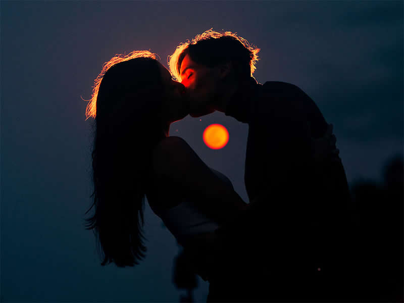 Cinematic silhouette of a couple kissing at dusk, backlit by a glowing orange sun, with their dark figures framed against a deep blue sky.