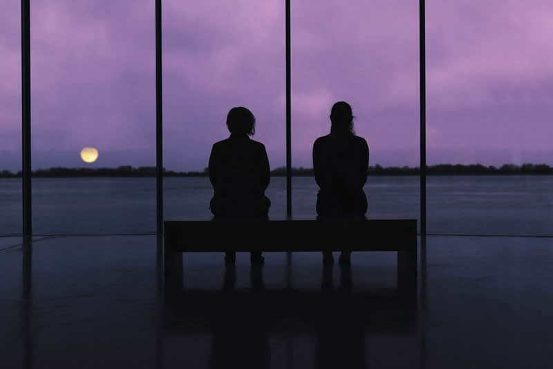 Cinematic photograph of two people sitting together on a bench, silhouetted against a purple twilight sky and calm water, with a glowing moon visible on the horizon.