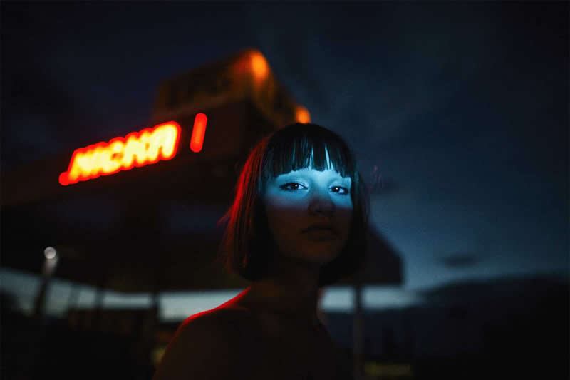 Cinematic nighttime portrait of a woman with short dark hair, illuminated by blue light and red neon reflections, with a glowing sign blurred in the background.