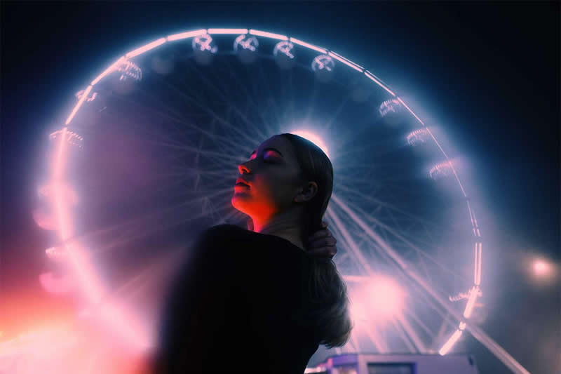 Cinematic nighttime portrait of a woman standing beneath a glowing Ferris wheel, illuminated by pink and red lights against a dark blue sky.