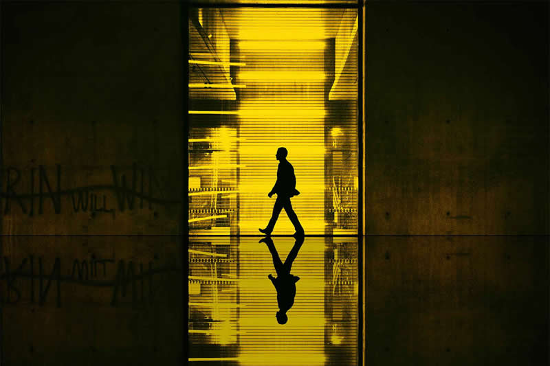 Cinematic photograph of a silhouetted person walking through a brightly illuminated yellow passage, with their reflection mirrored on the glossy floor and dark walls framing the scene.