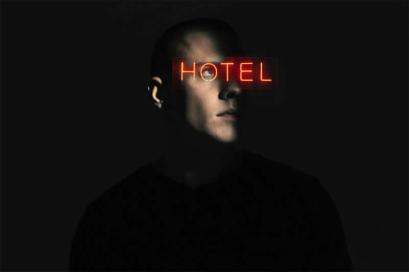 Cinematic portrait of a man in deep shadow with a glowing red &ldquo;HOTEL&rdquo; neon sign crossing his eyes against a dark background.