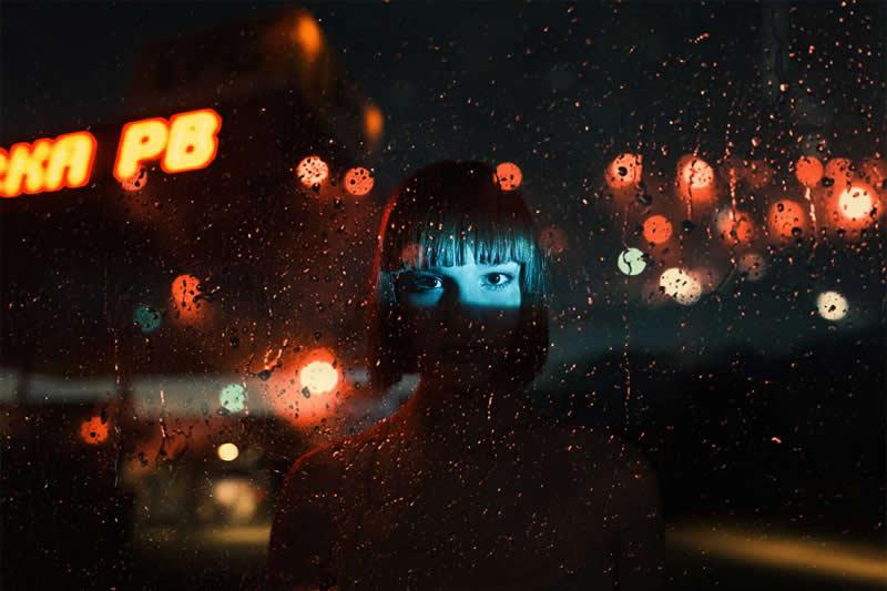 Cinematic nighttime portrait of a woman seen through a rain-covered window, with blue light illuminating her face and colorful orange and red city lights glowing behind her.