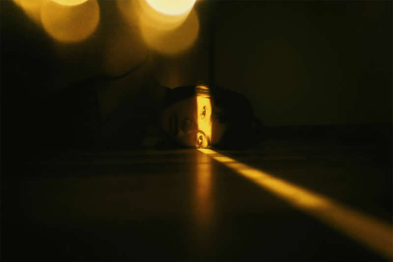 Atmospheric photograph of a dark interior with a narrow golden beam of light stretching across the floor toward a small illuminated opening, surrounded by deep shadows and warm bokeh.