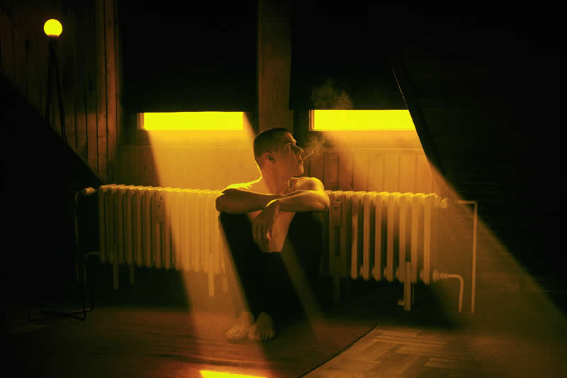 Cinematic portrait of a man sitting on the floor beside a radiator, surrounded by warm yellow window light, deep shadows, and atmospheric reflections in a dark room.