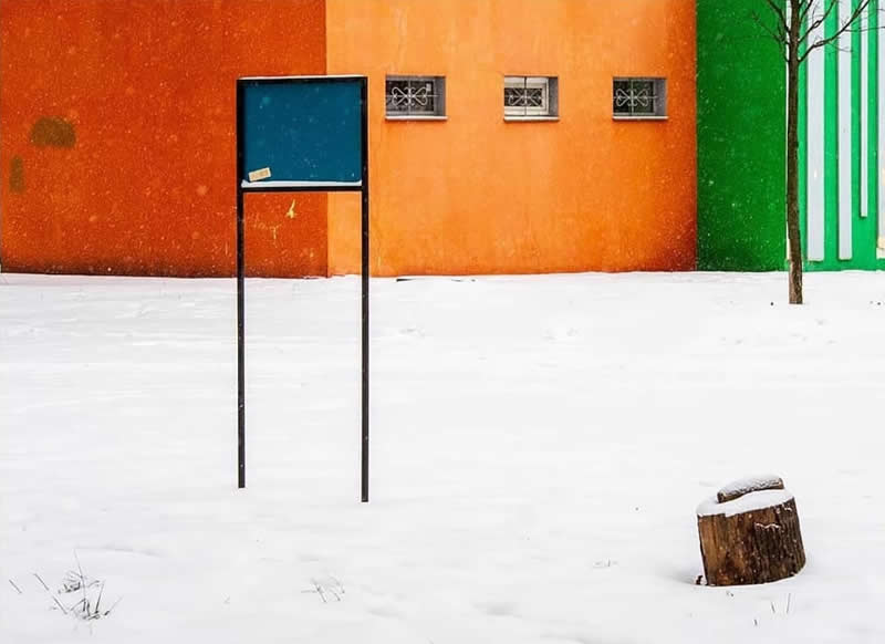 A snow-covered courtyard framed by vivid orange and green walls, with a freestanding sign, small tree, and snow-capped tree stump creating a minimalist composition.