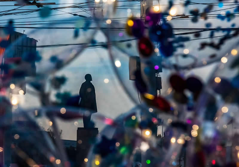 A silhouetted person standing in an urban setting, framed through blurred colorful lights, overhead wires, buildings, and glowing bokeh in the foreground.